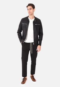 Giacca in pelle nera con tasche con zip e dettagli rigati, abbinata a una t-shirt bianca, pantaloni neri e sneakers marroni.