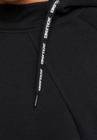 Sudadera negra con un cuello alto y tejido texturizado. Cuenta con un cordón y un logo de "JACK & JONES". Diseño minimalista sin patrones.