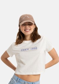 Balta trumpa T-marškiniai su mėlynu "ROXY 1990" tekstu. Pridėtas šviesiai rudas kepurė su tuo pačiu tekstu išsiuvinėtu balta spalva.