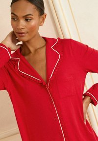 Rød pyjamasoverdel lavet af blødt stof med knaplukning foran, flæsekant langs kraven og ærmerne samt en lomme på venstre side.