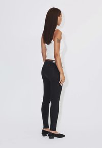Donna con capelli lunghi e scuri in piedi di lato che indossa una canotta bianca senza maniche, pantaloni neri aderenti e scarpe nere con tacco basso su sfondo bianco.