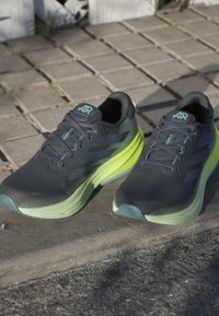 Hardloopschoenen met een zwarte mesh bovenkant, groene accenten en een gevoerde zool. Kenmerken zijn onder andere veters, een gevoerde kraag en gestructureerde patronen.