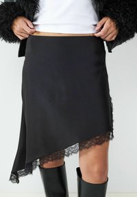 Schwarze asymmetrische Skirt aus strukturiertem Stoff, mit einer Spitzenborte am Saum, kombiniert mit hohen schwarzen Stiefeln.