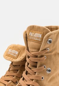 Stivali alta in tela beige con colletto imbottito, occhielli in metallo e lacci intrecciati. Presentano un'etichetta con "PALLADIUM" e una superficie testurizzata.