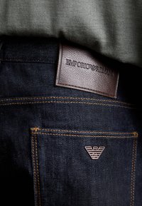 Mörkblå jeans i denim med en brun läderlapp präglad med "EMPORIO ARMANI", kombinerade med guldfärgade sömmar och en liten logodetalj på bakfickan.