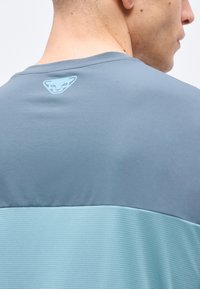 Άνδρας που φοράει δίχρωμο μπλε πουκάμισο με μικρό ανοιχτό μπλε λογότυπο πάνθηρα στο άνω πίσω μέρος κοντά στον λαιμό.