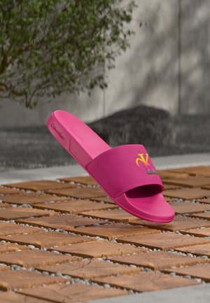 Sandalia tipo slide Calvin Klein de color rosa intenso con correa de tela que presenta el logo multicolor CK, mostrada al aire libre sobre adoquines de madera con vegetación y fondo de piedra.