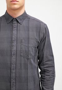 Mężczyzna ubrany w ciemnoszarą, zapinaną na guziki koszulę w kratę z kieszenią na piersi, pokazany od środkowej części szyi do talii na jednolitym tle.