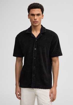 Junger Mann mit lockigem Haar, der ein schwarzes, kurzärmeliges, strukturiertes Hemd mit Knöpfen und weiße Hosen trägt, stehend vor einem einfarbigen Hintergrund.