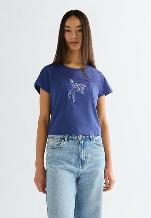 Młoda kobieta z długimi ciemnymi włosami, ubrana w niebieską koszulkę z białym konturem jeźdźca rodeo na kozłującym koniu oraz jasnoniebieskie dżinsy.