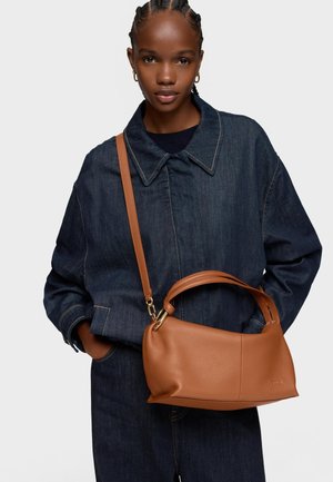 Donna con giacca e pantaloni di denim scuro, che porta una borsa in pelle marrone medio con tracolla e manici superiori.