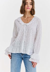 Kobieta ubrana w białą teksturowaną bluzkę z marszczonymi mankietami, dołem i kołnierzem, w połączeniu z jasnoniebieskimi dżinsami i złotym naszyjnikiem z zawieszką.