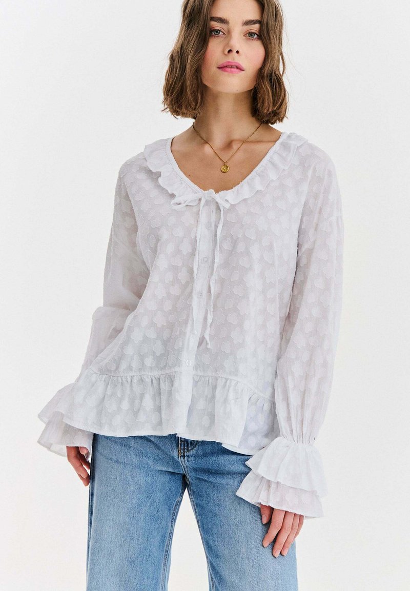 Kobieta ubrana w białą teksturowaną bluzkę z marszczonymi mankietami, dołem i kołnierzem, w połączeniu z jasnoniebieskimi dżinsami i złotym naszyjnikiem z zawieszką.