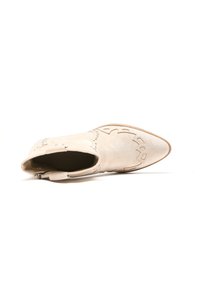 Botín de ante beige hasta el tobillo con punta afilada, costuras decorativas y acentos metálicos en la parte superior. Textura suave y diseño curvado.