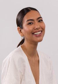 Frau mit glattem, dunklem Haar in einem tiefen Pferdeschwanz, die ein helles Leinenoberteil und goldene Creolen trägt und vor einem schlichten Hintergrund lächelt.