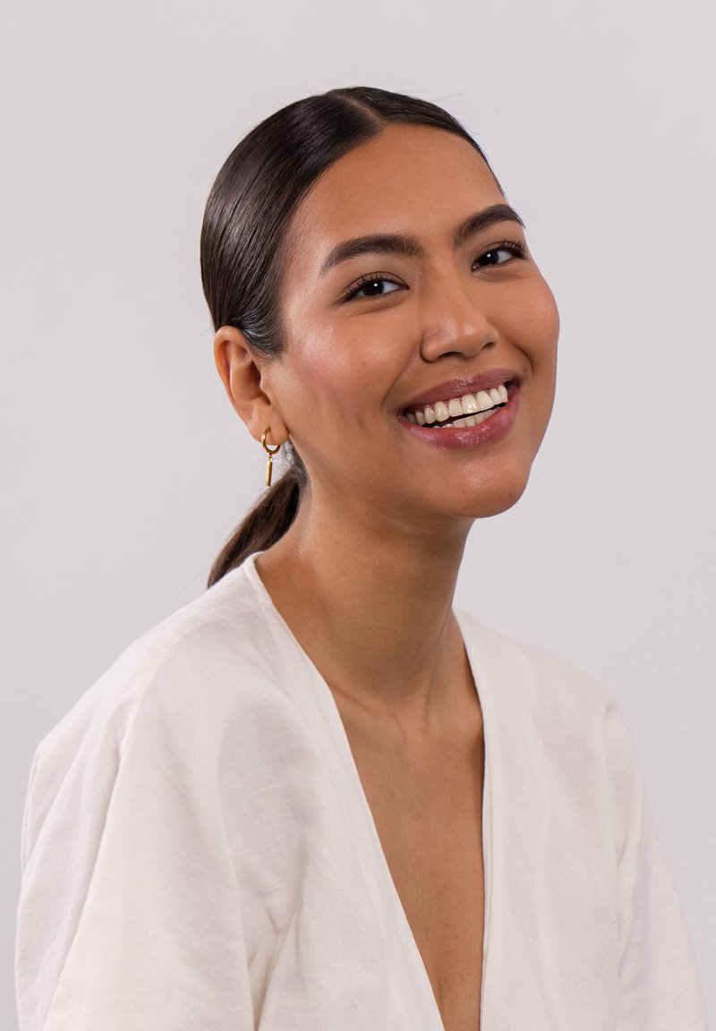 Frau mit glattem, dunklem Haar in einem tiefen Pferdeschwanz, die ein helles Leinenoberteil und goldene Creolen trägt und vor einem schlichten Hintergrund lächelt.