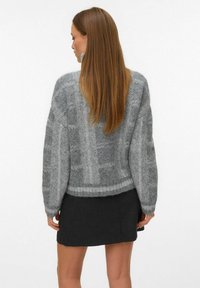 Sieviete ar gariem taisniem matiem, kas valkā pelēku adītu džemperi ar rakstu un īsu tumšu svārku, vērsta prom no kamerām.