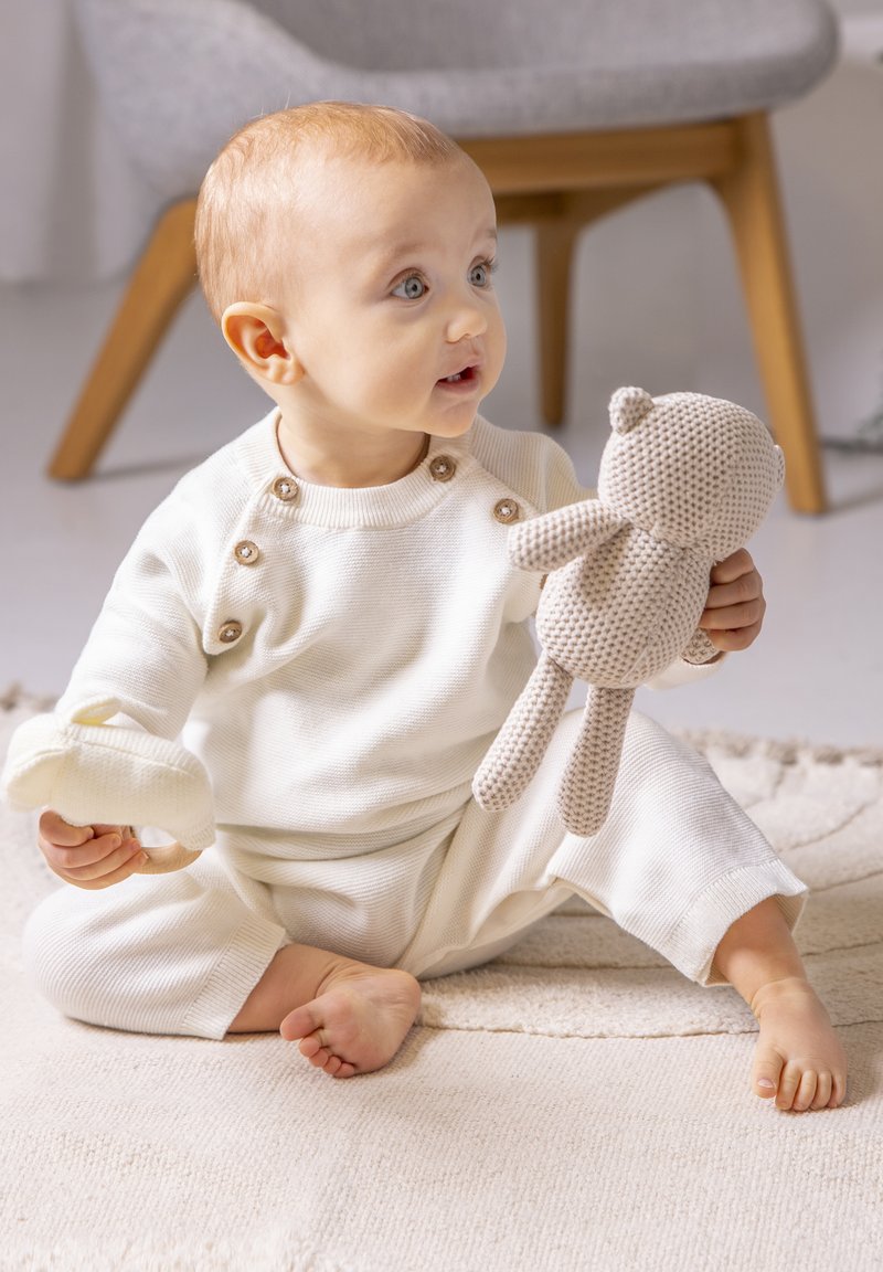 Crème-kleurig zacht katoenen outfit met drukknoopjes, kind dat een gebreide pluche beer en een klein crèmekleurig speeltje vasthoudt, zittend op een gestructureerd kleed.