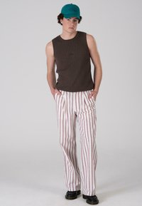 Ribbed rjav brezrokavnik v kombinaciji z belimi hlačami s črtami v rdeči in bež barvi. Manekenka nosi turkizno kapo in črne masivne škornje.