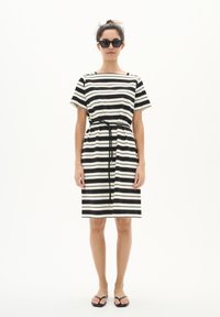 Abito a righe in bianco e nero, caratterizzato da una vestibilità rilassata, maniche corte e una cintura da annodare in vita. Indossato con occhiali da sole neri e sandali.