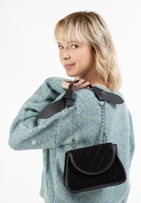 Sort quiltet håndtaske med en struktureret form, sølvfarvet kædestrop og læderdetaljer, båret over en lyseblå sweater.