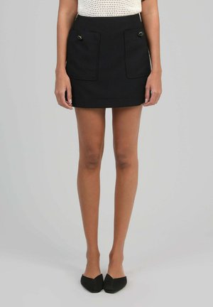 Femme portant une mini-jupe noire avec deux poches avant et des ballerines noires pointues, se tenant devant un fond gris uni.