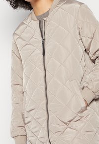 Quiltad bomberjacka i ljusbeige, tillverkad av mjukt material. Har en framdragkedja, ribbade ärmslut och sidofickor med ett diamantmönster.