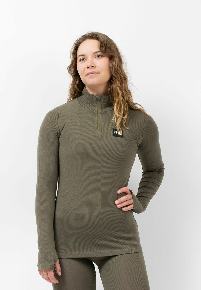 Femme aux cheveux longs et ondulés, portant un haut à manches longues vert olive et un legging assorti, se tenant avec une main sur la hanche contre un fond blanc.