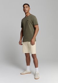Olijfgroene katoenen T-shirt met korte mouwen en een ronde hals, gecombineerd met crèmekleurige shorts. Schoenen zijn lichtgrijs met beige accenten.