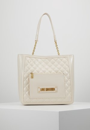Kremfarvet quiltet tote-taske med gulddetaljer, der har en lynlåslomme foran og logo-plade. Glat kunstlædermateriale, struktureret form.