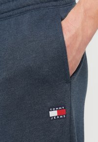 Mână în buzunarul pantalonilor de trening gri închis, cu un patch cu logo-ul Tommy Jeans aproape de marginea buzunarului.