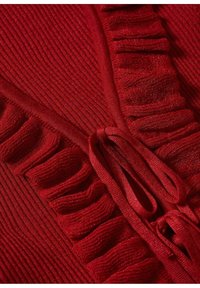 Rotes gestricktes Kleidungsstück mit gerippter Textur, das Rüschen entlang des Randes und eine Bindeschließe aufweist, wobei komplizierte Details und Tiefen zur Geltung kommen.