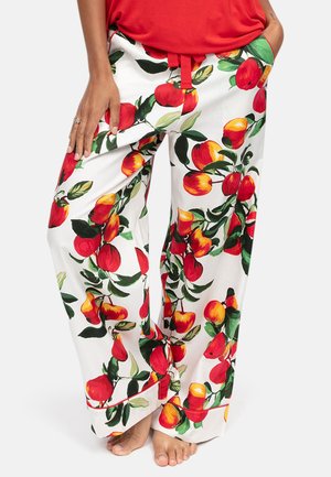 Vrouw draagt witte broek met wijde pijpen met rode en gele fruit- en groene blaadjesprint, rood topje, staat blootsvoets met één hand in de zak.