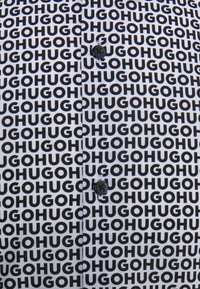 Camisa de manga corta blanca con un patrón de texto negro repetido "GOHUGO". Presenta un cierre de botón y acentos de botón negro.