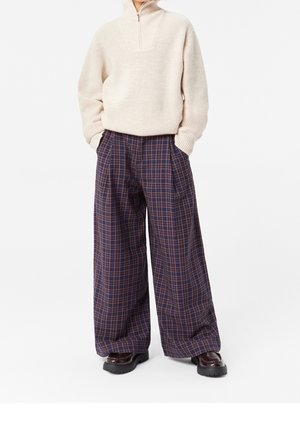 Beiger Pullover mit Reißverschluss am Kragen und geripptem Muster, kombiniert mit weit geschnittenen marineblauen karierten Hosen mit braunen und rostfarbenen Linien. Schwarze Schuhe mit dicker Sohle.