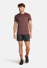 Bordeauxfarbendes T-Shirt mit kurzen Ärmeln, auf dem eine horizontale Linienzeichnung eines Zeltes und eines Feuers zu sehen ist. Getragen zu einer schwarzen Shorts, orangefarbigen Socken und schwarzen Schuhen.
