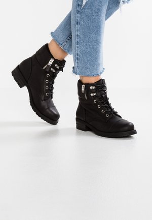 Feet wearing black leather lace-up ankle boots with side zippers and light blue rolled-up frayed hem jeans on white floor.