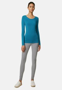 Långärmad turkos topp med rundad halsringning, kombinerad med grå leggings. Leggingsen har en slät textur. Modellen bär ljusa skor.