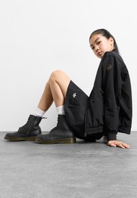 Chaqueta bomber negra con un parche de logo, combinada con una falda negra. Botas gruesas negras con cordones y suela amarilla, acompañadas de calcetines blancos hasta el tobillo.