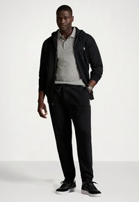 Sweatshirt preta com fecho de correr, com camisa polo cinzenta por baixo, combinada com calças de treino pretas e ténis pretos. Texturas suaves e corte descontraído.