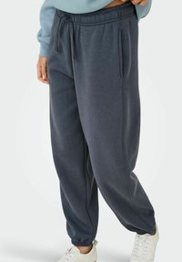 Pantaloni della tuta grigio scuro realizzati in tessuto morbido, con cintura elastica, tasche laterali e polsini a taper. Indossati con sneakers chiare.