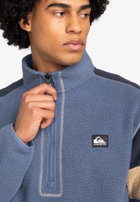 Polarowy rozpinany pulower w kolorze niebieskim z ćwierć zamkiem, kontrastującymi ciemnoniebieskimi i kremowymi akcentami oraz naszywką z logo na piersi. Miękka tekstura.