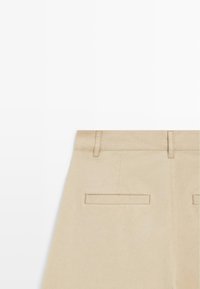 Beżowa spódnica pokazująca górną część z tyłu z szlufkami na pasek i dwoma poziomymi kieszeniami w listwach, na jednolitym białym tle.