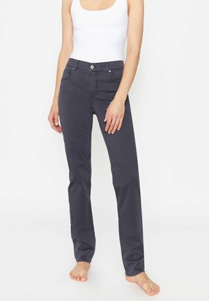 Personne portant un pantalon droit gris foncé et un haut blanc sans manches, debout pieds nus sur un fond blanc.