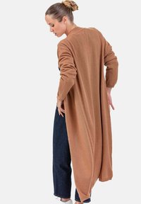 Langer, brauner Cardigan aus weichem, strukturiertem Stoff mit lockerem Sitz. Verfügt über lange Ärmel und einen Rückenschlitz, kombiniert mit dunkelblauen Jeans und weißen Sneakern.