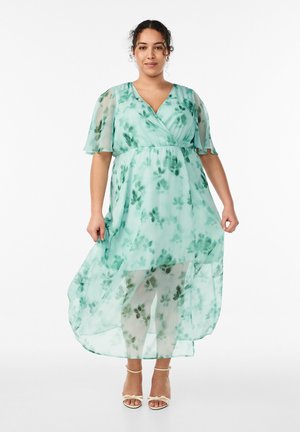 Žena u lepršavoj, svijetlozelenoj haljini s cvjetnim uzorkom, prozirnim kratkim rukavima i sandalama s remenčićima oko gležnjeva, stoji nasuprot bijele pozadine.