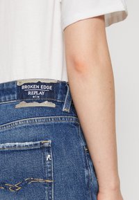 Farkut denim sinisessä pesussa, joissa on teksturoitu pinta, takataskut ja vyötärölle ommeltu merkki, jossa lukee "Broken Edge REPLAY".