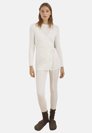 White, long-sleeve wrap top with tie detail, paired with light-colored leggings and brown, fuzzy slip-on slippers. Smooth texture, minimal design.