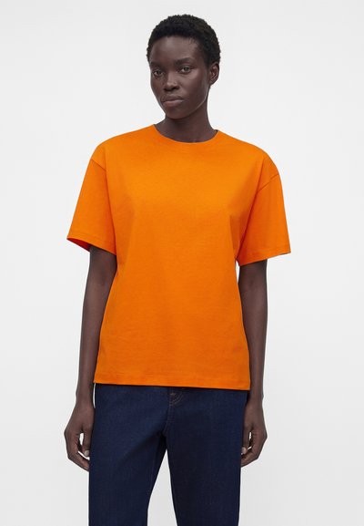 Γυναίκα με κοντά μαλλιά που φοράει απλό φωτεινό πορτοκαλί κοντομάνικο μπλουζάκι και σκούρο μπλε τζιν, με λευκό φόντο.