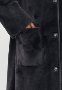 Manteau en fausse fourrure doux de couleur gris foncé, doté d'un bouton de fermeture et de poches latérales. Texture lisse avec une coupe décontractée et des manches longues.